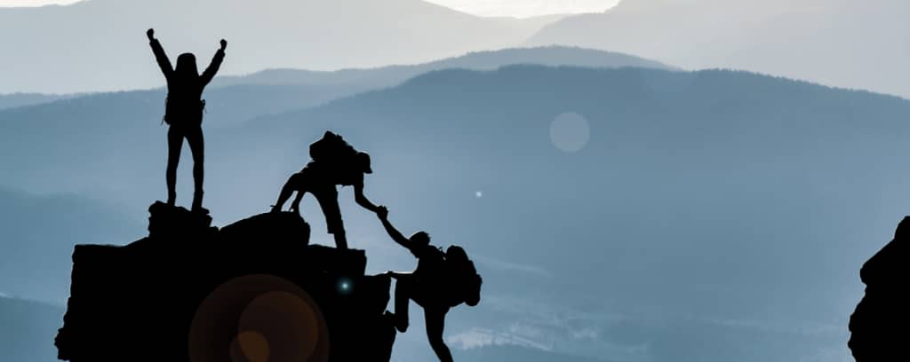 Photo of three people reaching a peak; one is helping another