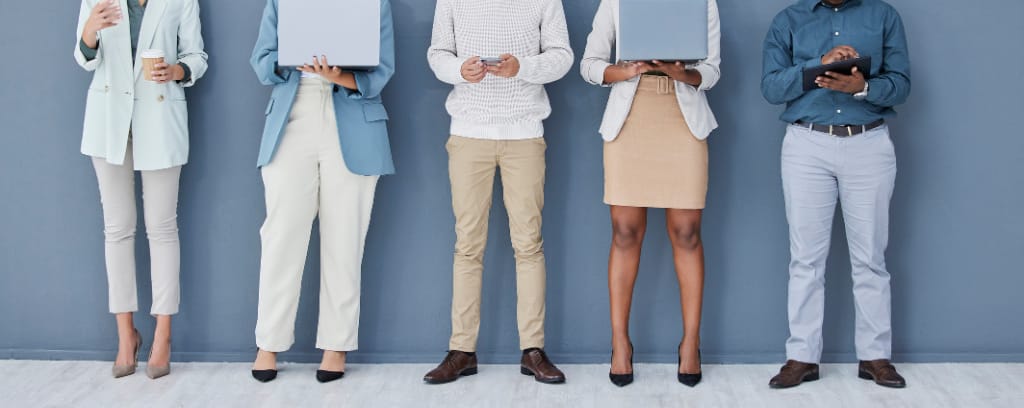 Five job candidates diverse in age, gender, style, resume medium, and more, standing against a blue-gray wall, waiting for their job interviews.