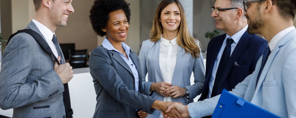 Group of employees wearing suits and smiling while two shake hands.