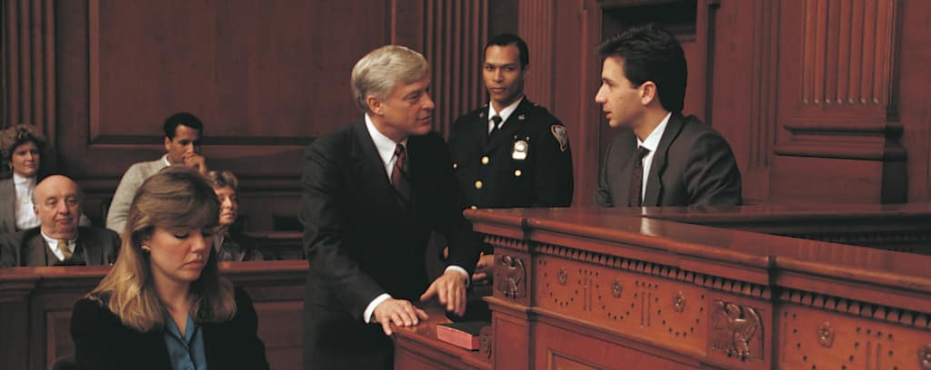 Photo of attorney questioning a witness in a courtroom with a bailiff in the background and court reporter in the foreground