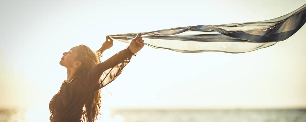 Photo of woman on the beach enjoying the warmth on her face and wind blowing a light scarf she holds overhead